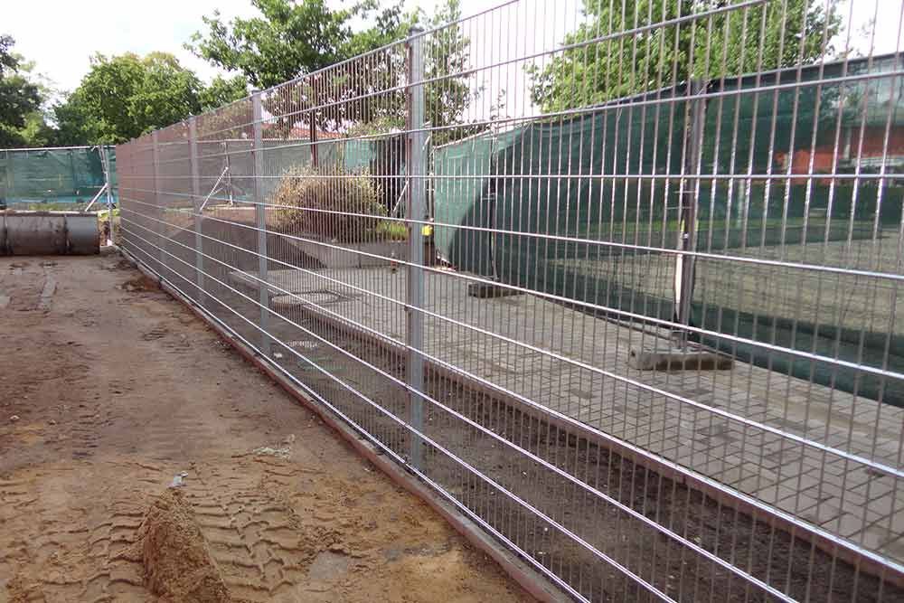 Ein Metallzaun umgibt eine unbefestigte Fläche mit Bäumen im Hintergrund