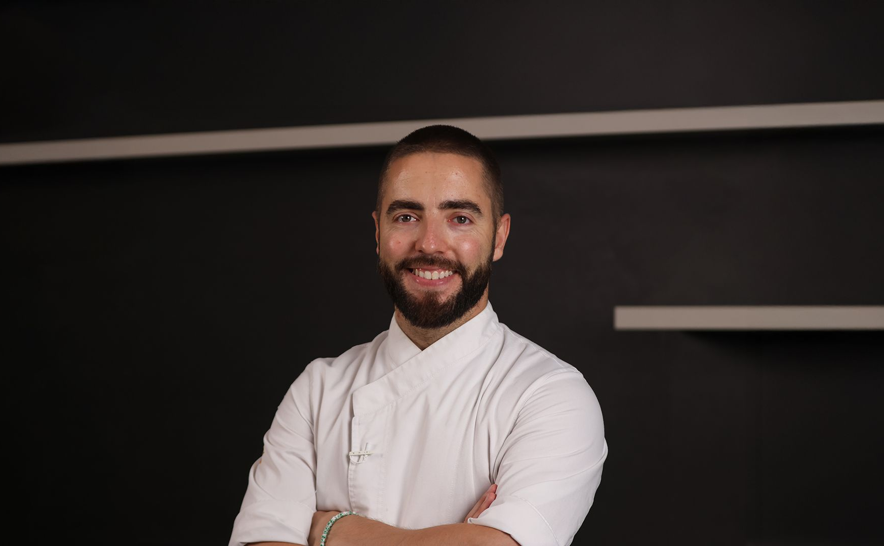 Chef con barba y chaqueta blanca, con los brazos cruzados, sonriendo a la cámara en un entorno moderno.