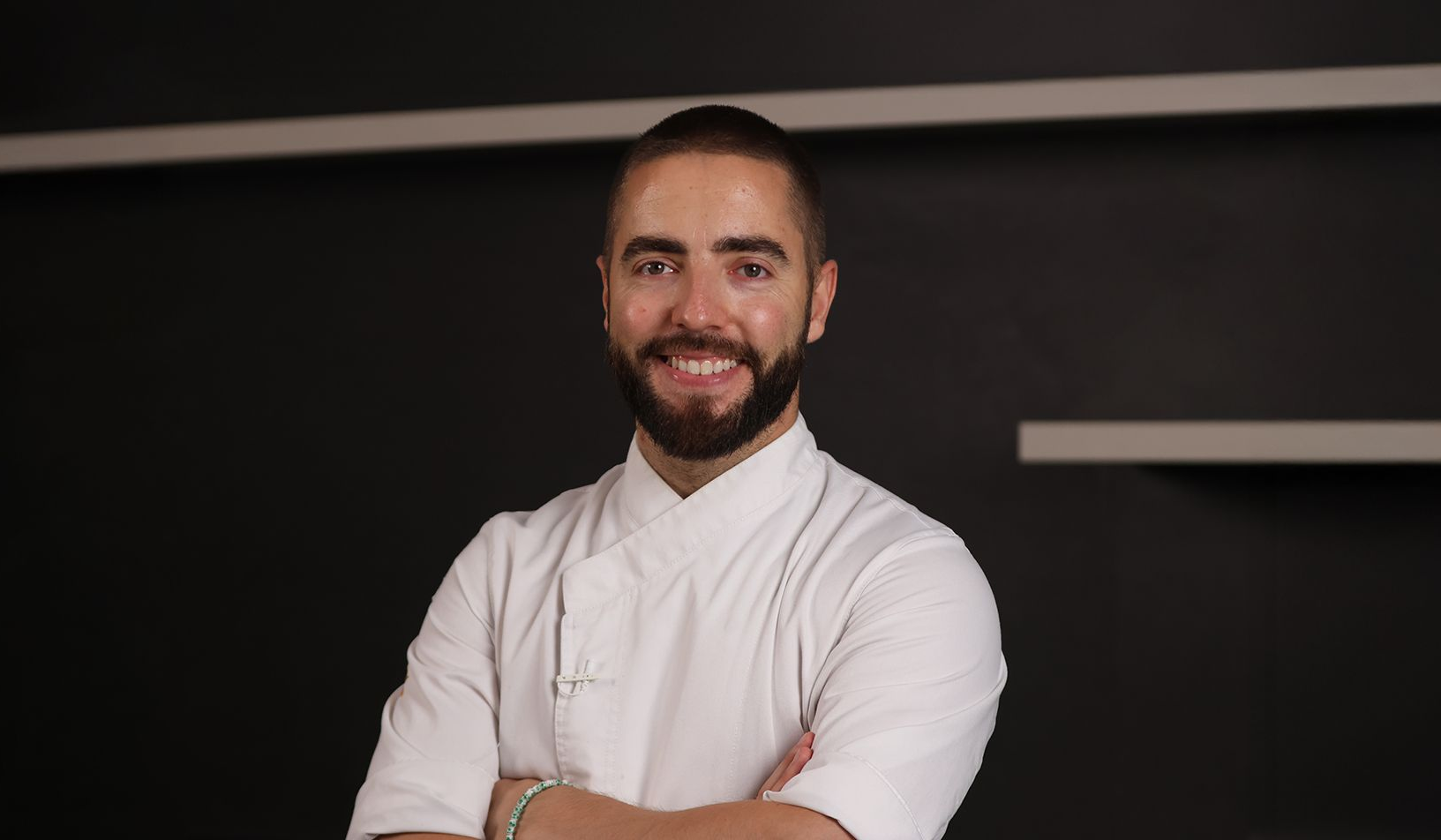 Chef con barba y chaqueta blanca, con los brazos cruzados, sonriendo a la cámara en un entorno moderno.