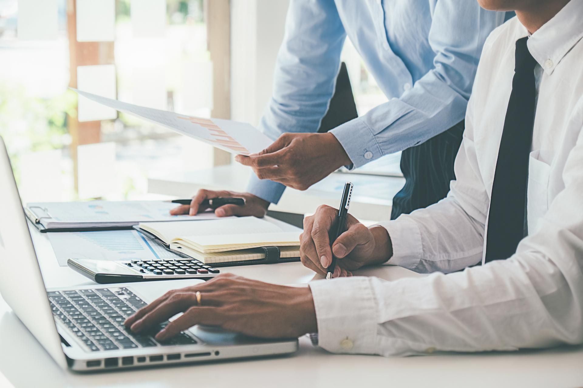 Ein Mann tippt auf einem Laptop, während ein anderer Mann auf ein Blatt Papier schaut.