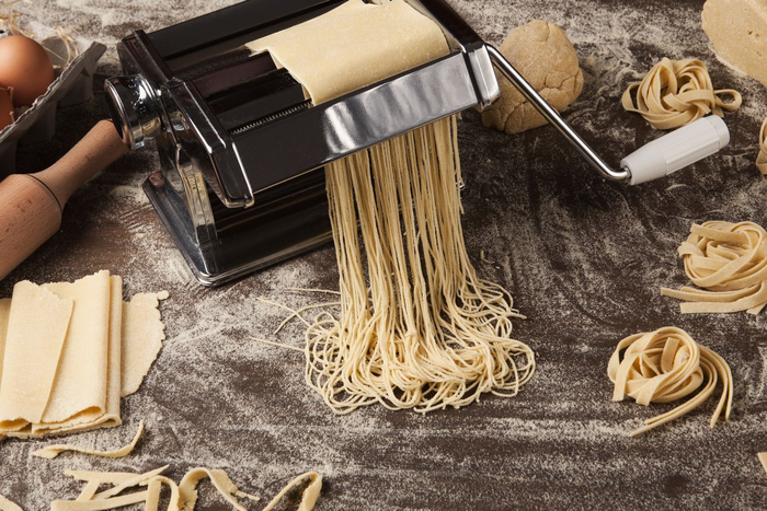 Une machine à pâtes découpe une feuille de pâte fraîche en nouilles sur une surface farinée, avec plusieurs moules à pâtes à proximité.