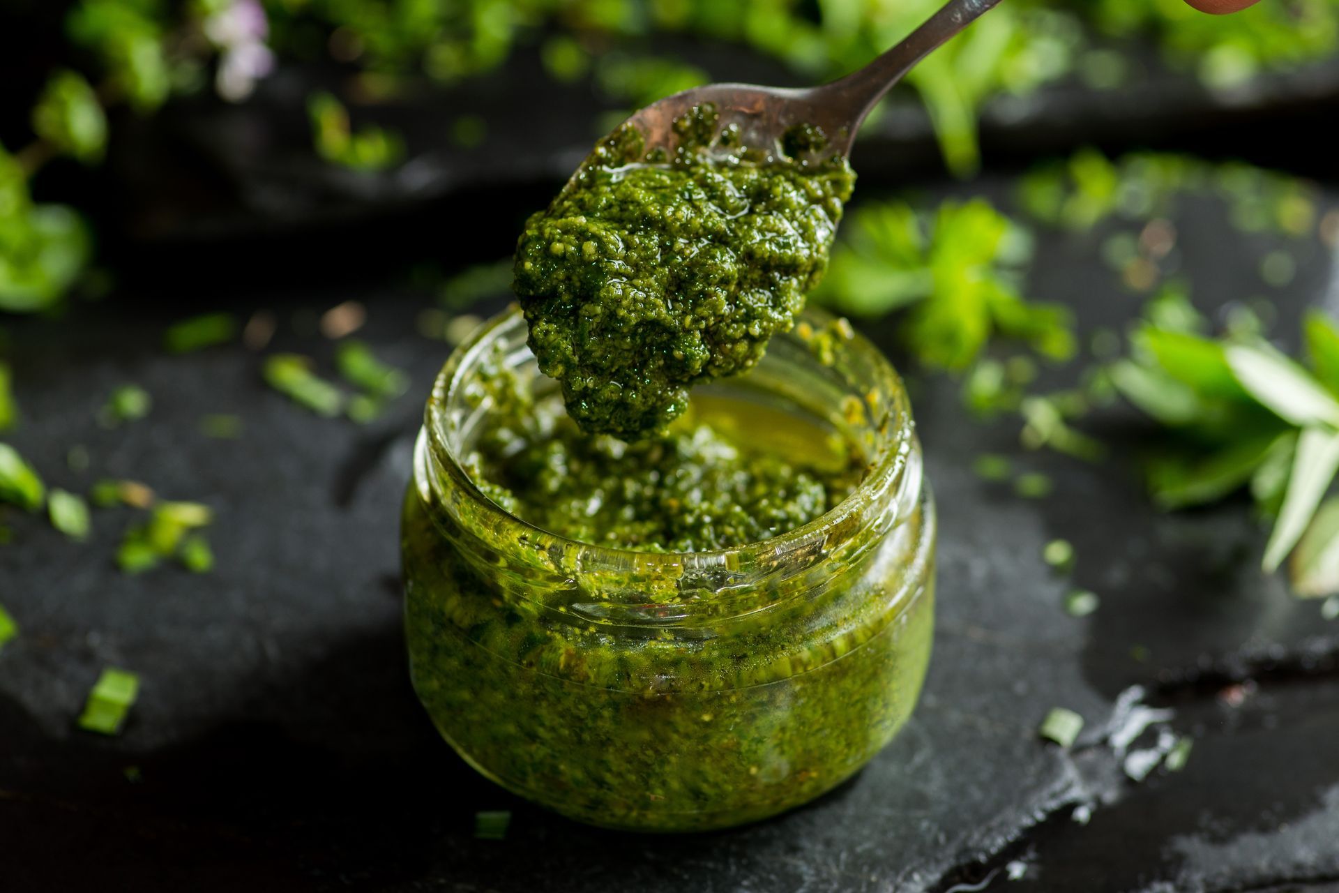 Une cuillère en argent prélève une cuillerée de pesto d'un vert éclatant dans un bocal en verre posé sur une surface d'ardoise sombre et rustique.