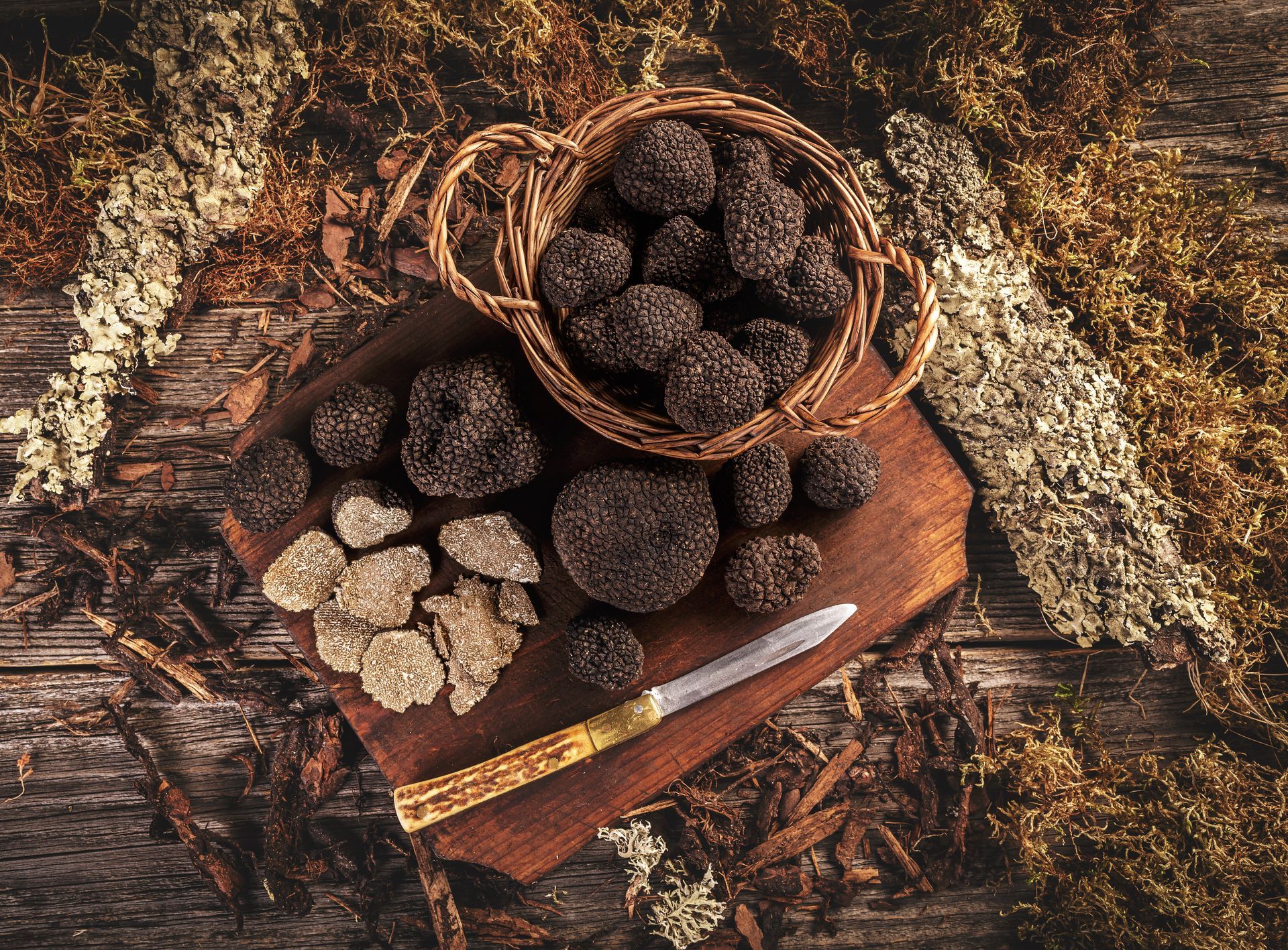 Truffes noires dans un petit panier en osier et posées en vrac sur une planche en bois avec un couteau d'office, le tout sur un fond rustique de mousse.