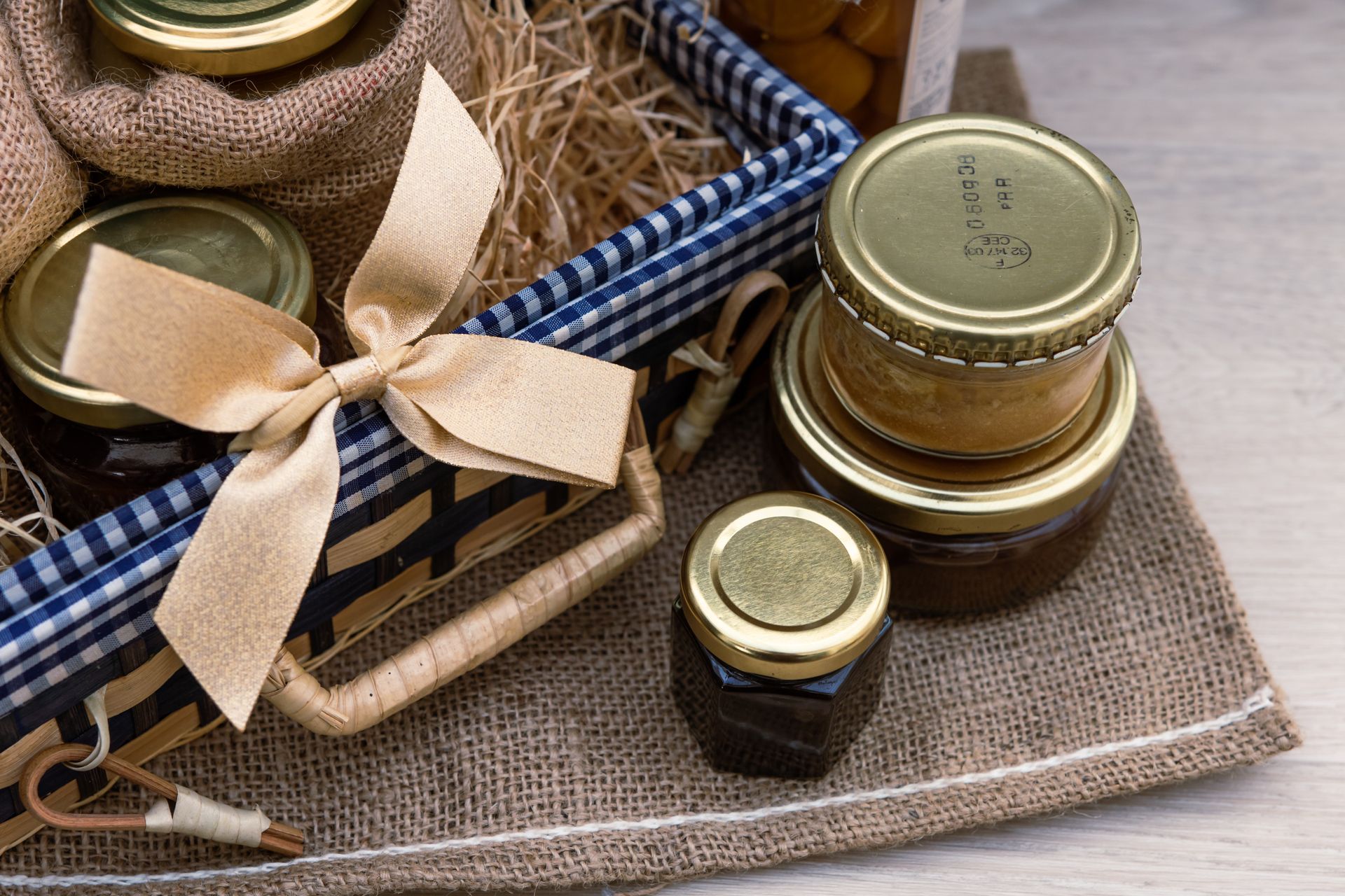 Petits bocaux en verre avec couvercles dorés et ruban beige, nichés dans un panier doublé de toile de jute à bordure vichy bleue et blanche.