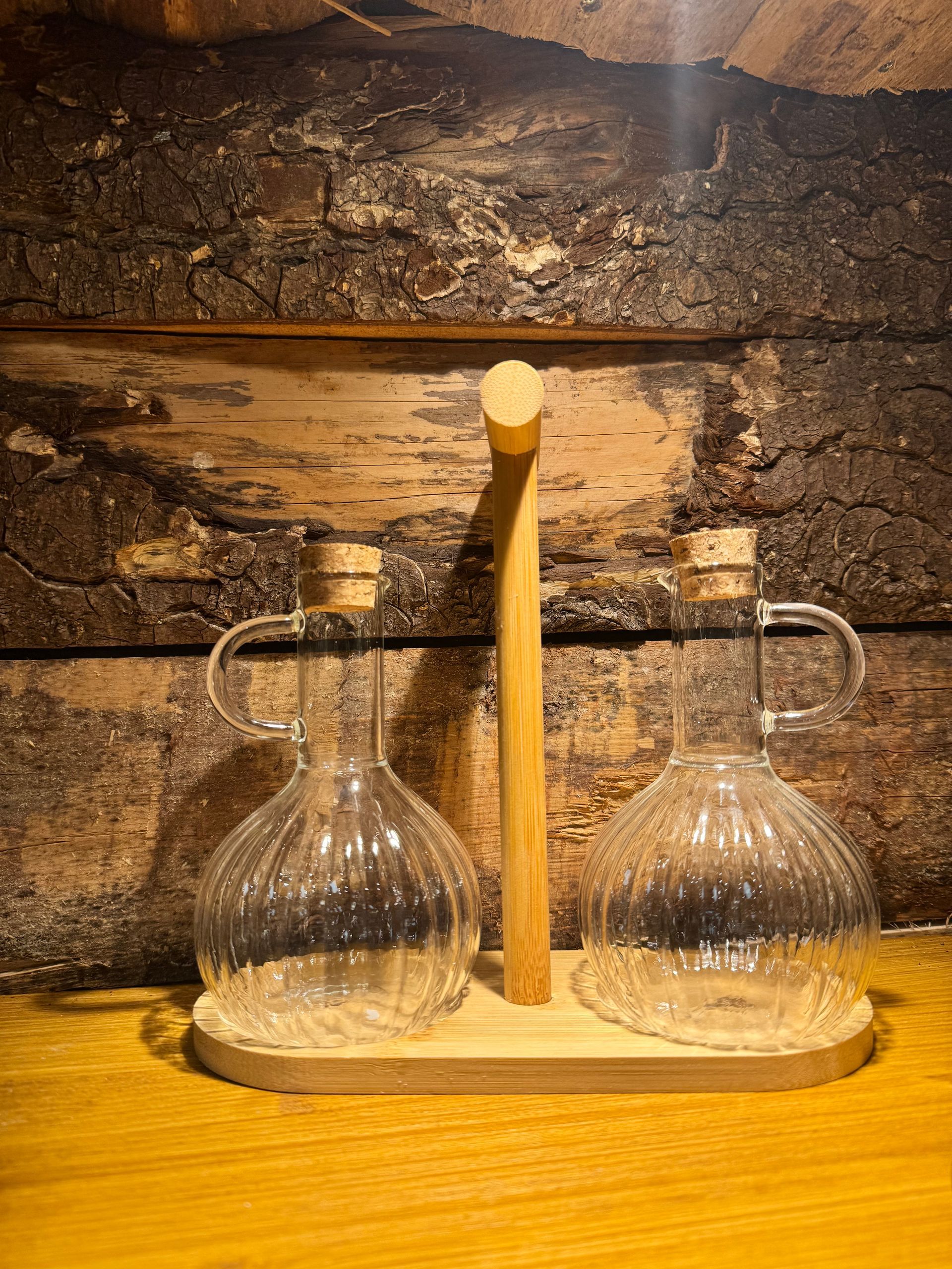 Deux burettes à huile et à vinaigre en verre côtelé, avec bouchons de liège, posées sur un support en bois contre un mur rustique en planches de bois.