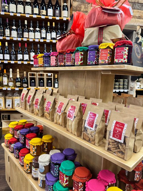 Des étagères dans un magasin présentent des bouteilles de vin, des bocaux assortis avec des couvercles colorés et des sacs de produits alimentaires.