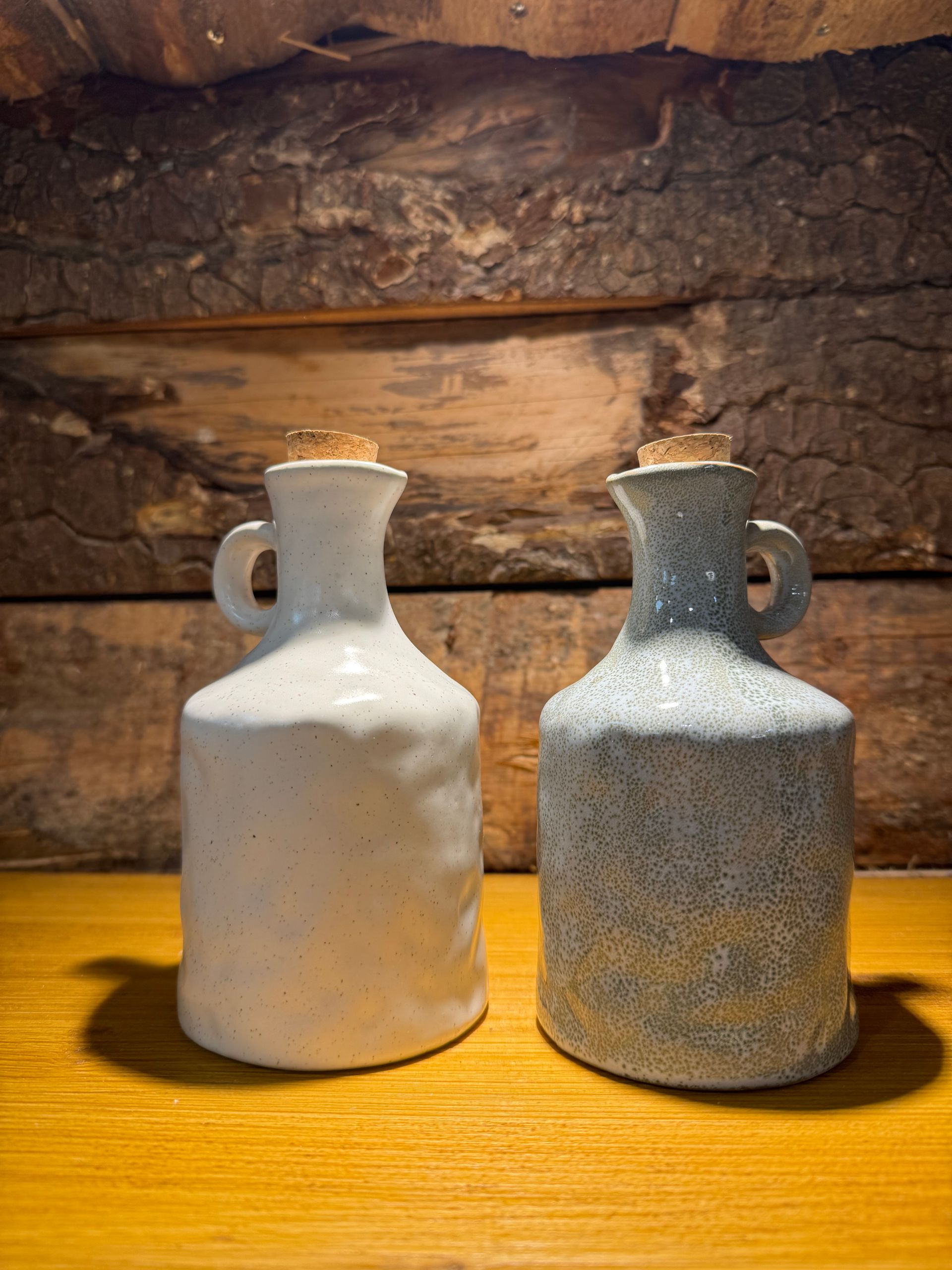 Deux bouteilles, en céramique, artisanales avec bouchons de liège, l'une blanche et l'autre à pois bleus, sont posées côte à côte sur une surface en bois.