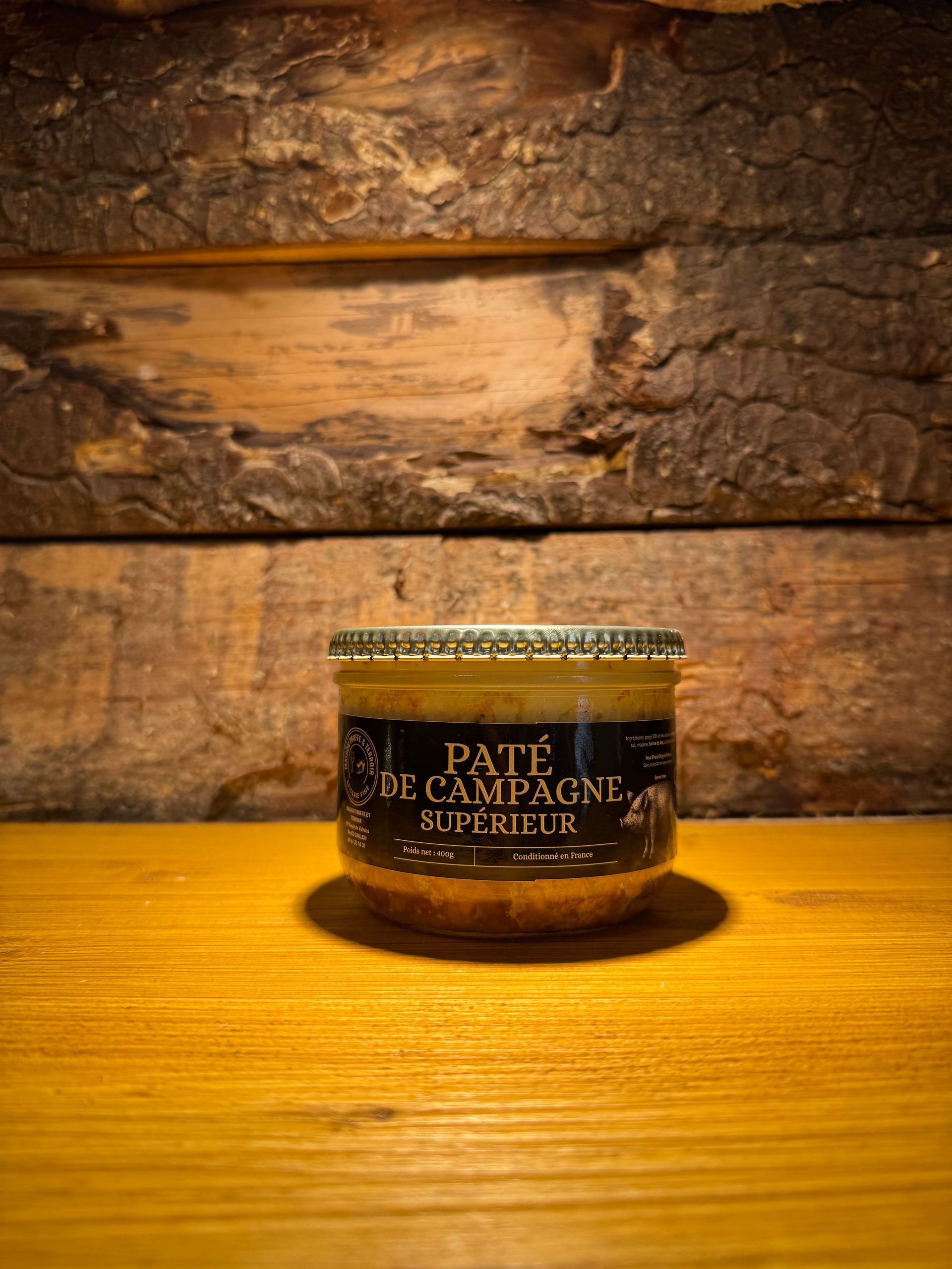 Un pot de pâté de campagne repose sur une surface en bois contre un mur rustique en planches de bois, éclairé par une lumière chaude.