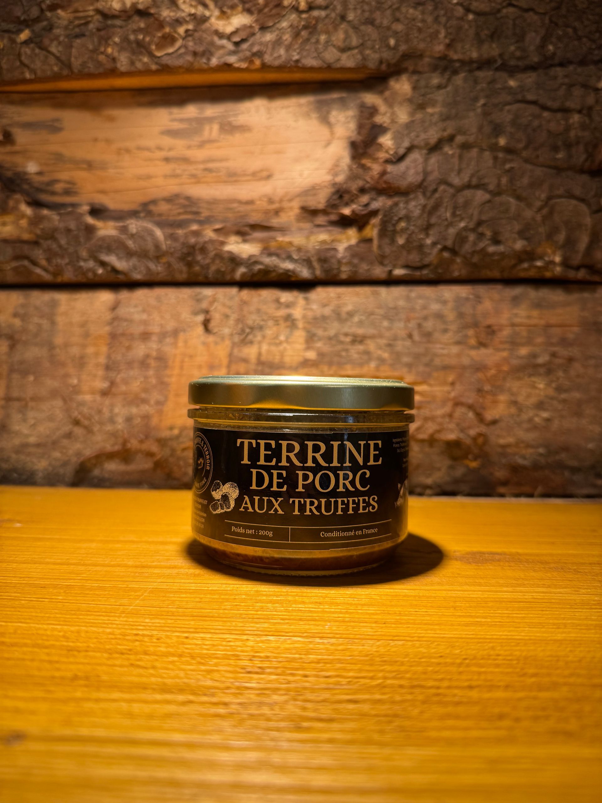 Un petit bocal en verre étiqueté « Terrine de Porc aux Fruits » est posé sur une surface en bois devant un fond rustique en planches de bois.