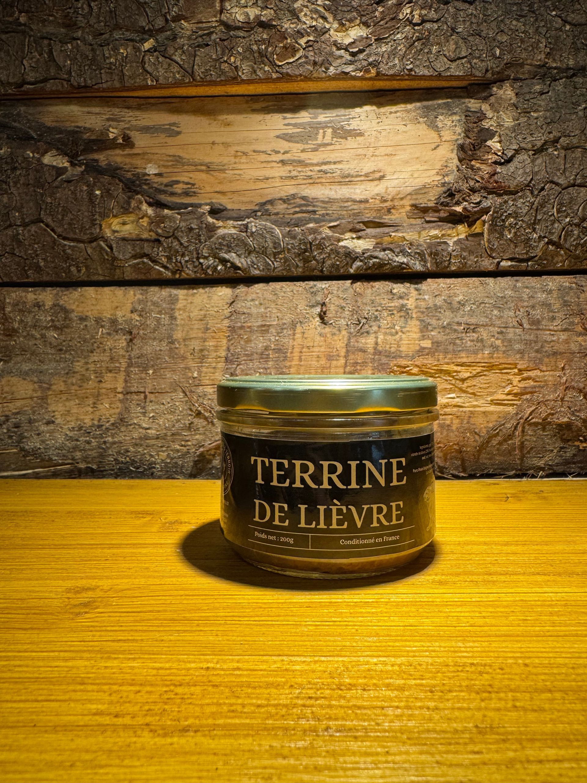Un pot de terrine de lièvre repose sur une surface en bois, devant un fond rustique recouvert d'écorce sombre.