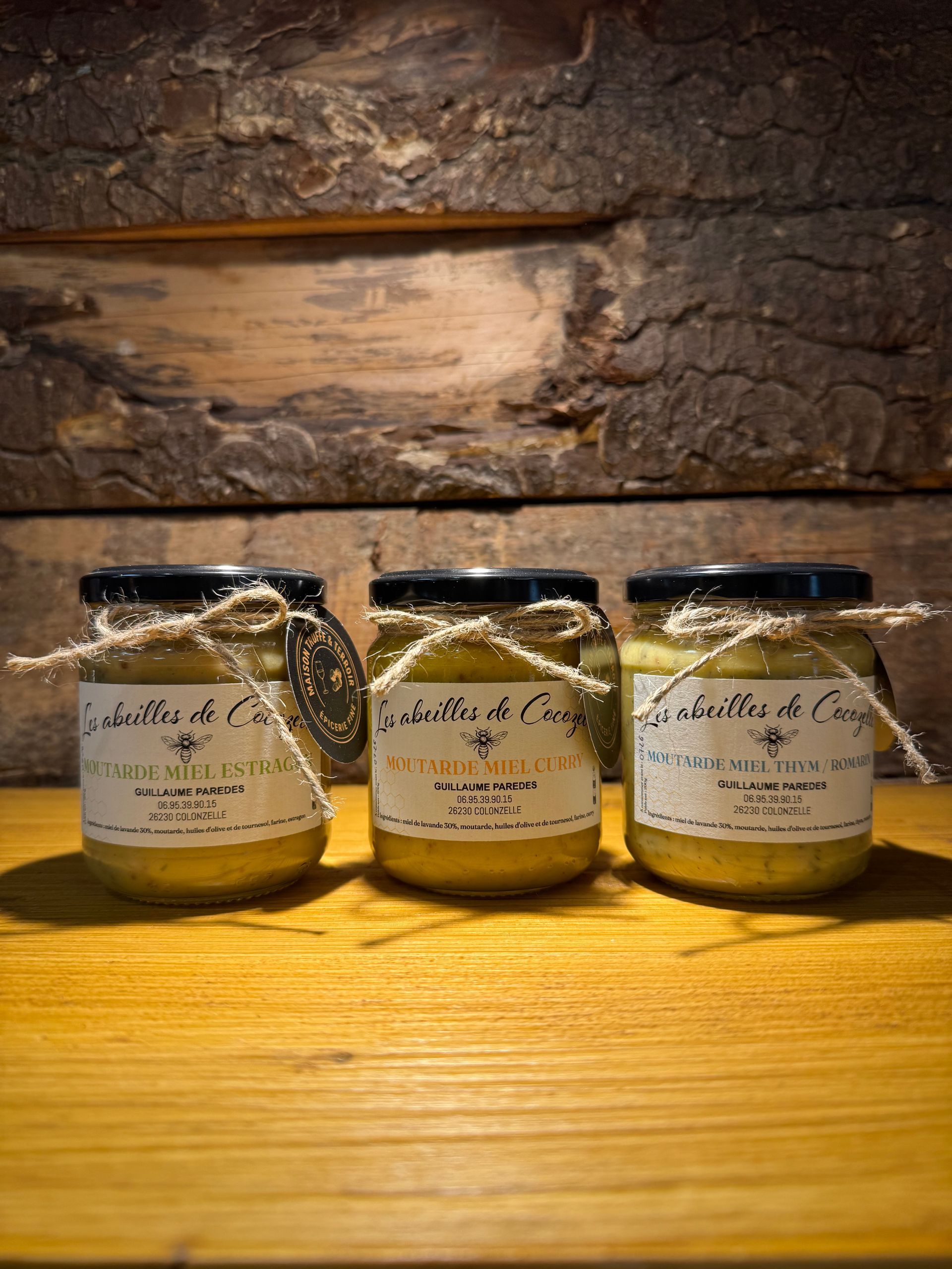 Trois pots de miel, étiquetés avec des nœuds en ficelle rustique, alignés sur une surface en bois devant un fond de planches de bois sombres.