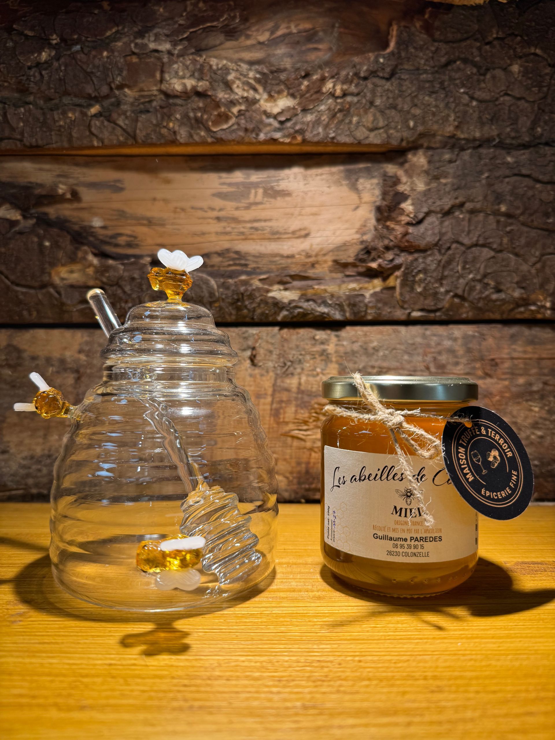 Un pot à miel en verre en forme de ruche, avec une louche, posé à côté d'un pot de miel sur une surface en bois, devant un fond de rondins rustiques.