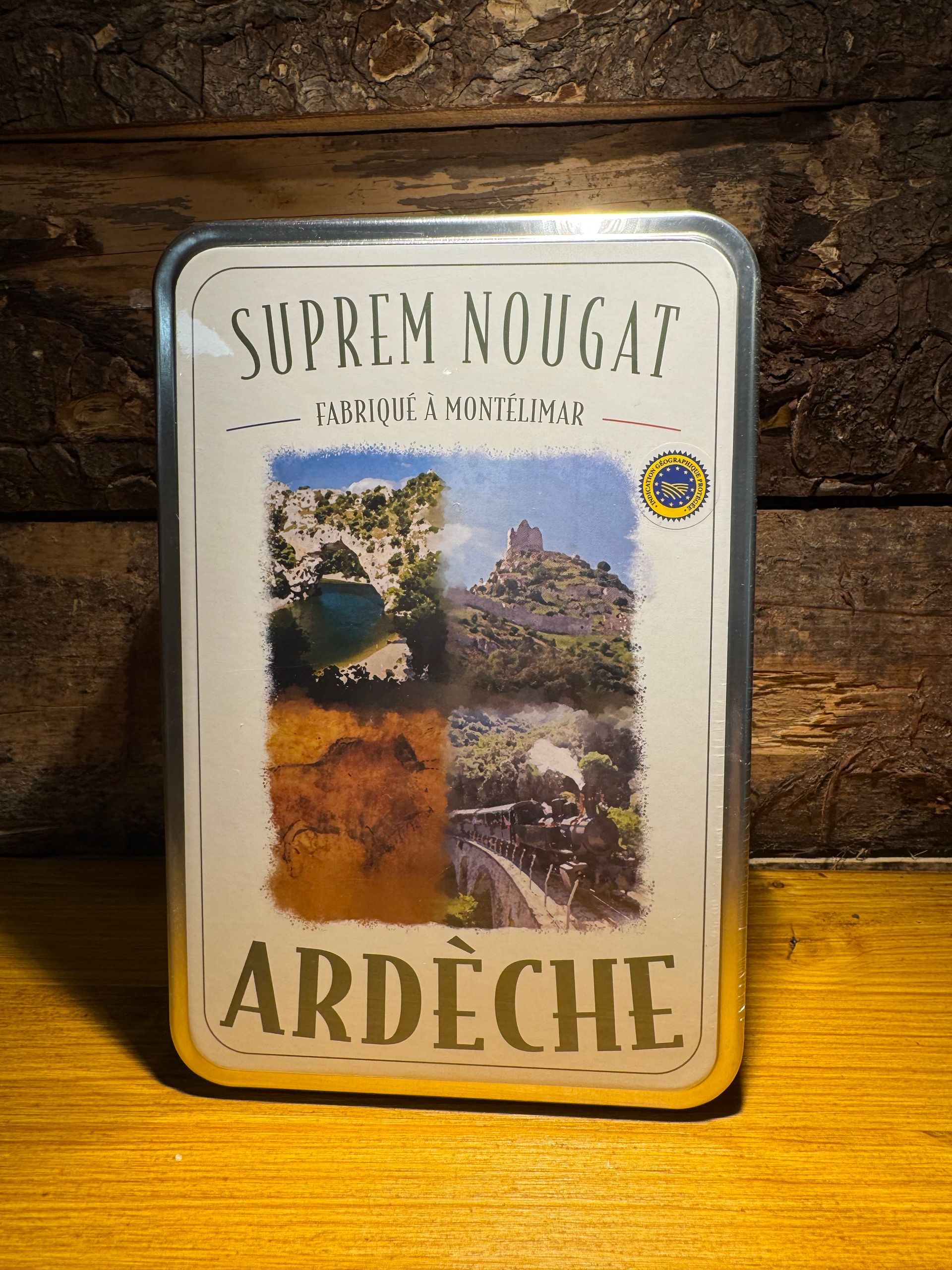 Une boîte de « Suprême Nougat » d'Ardèche, ornée d'un collage de photos de paysages, posée sur une surface en bois.