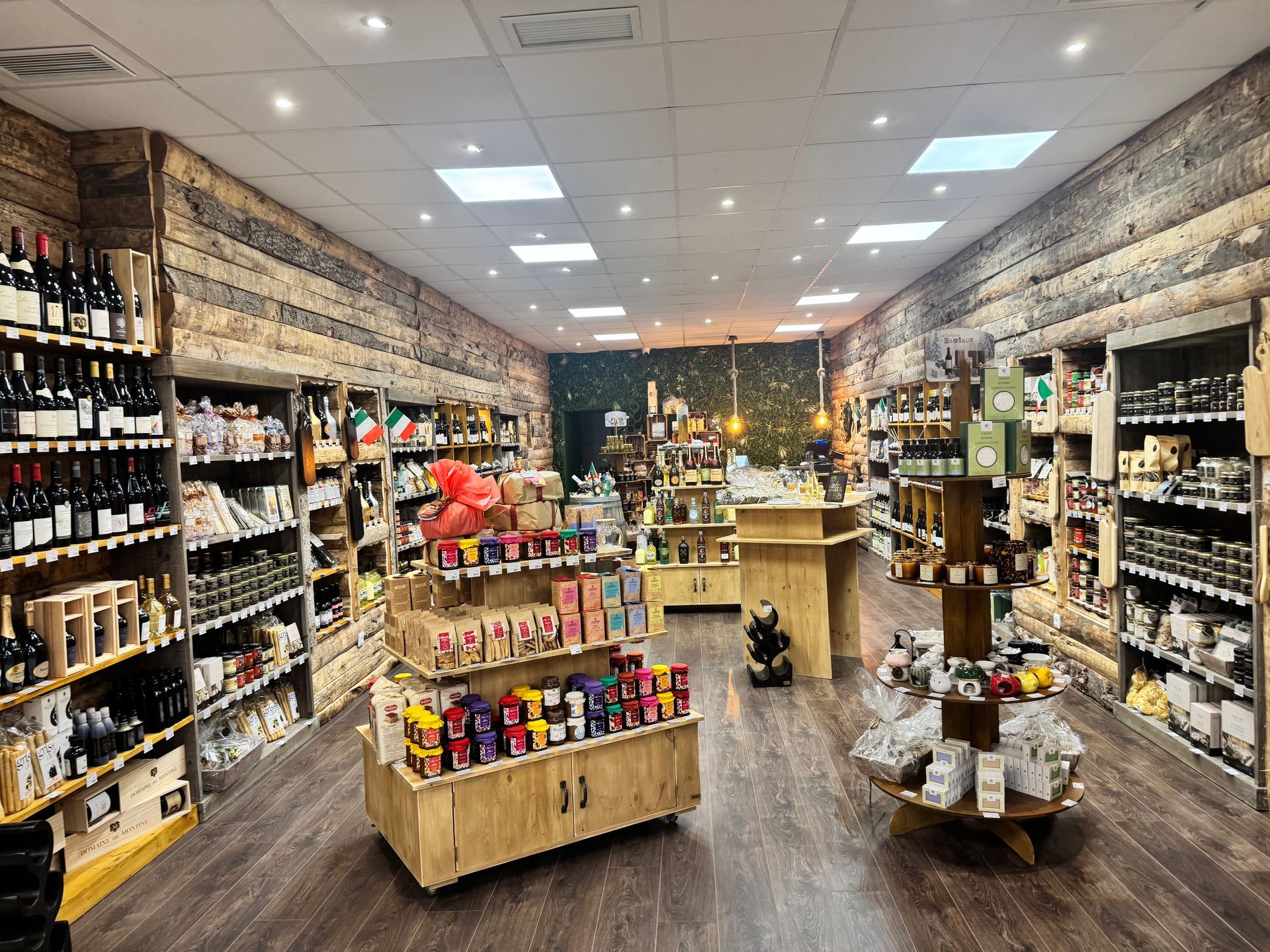 Vue intérieure d'une épicerie rustique avec des murs lambrissés, des étagères en bois remplies de vin et des présentoirs centraux pour les produits.