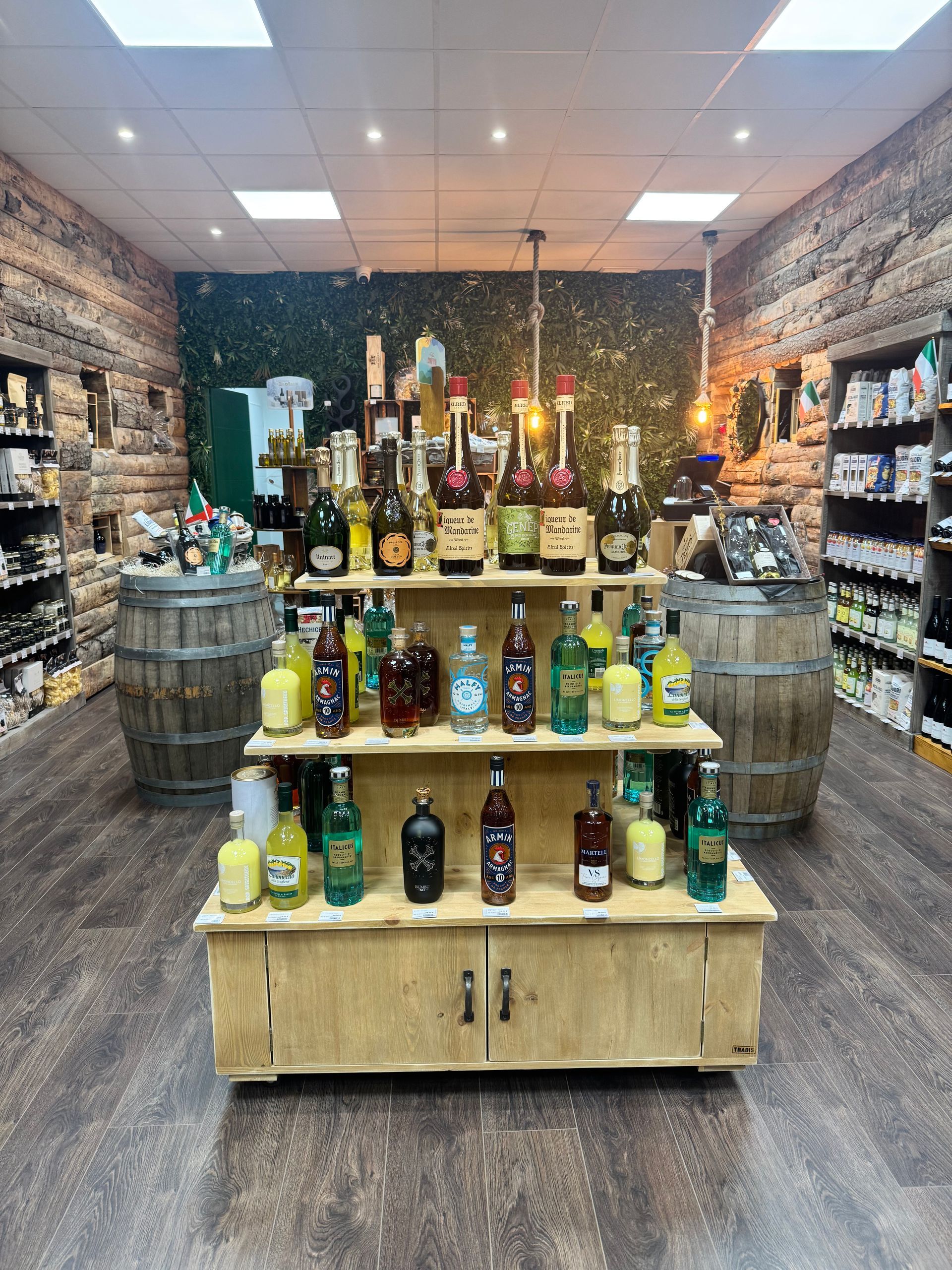 Dans un magasin, un présentoir en bois rempli de bouteilles d'alcool en verre de différentes tailles est flanqué de deux grands tonneaux en bois.
