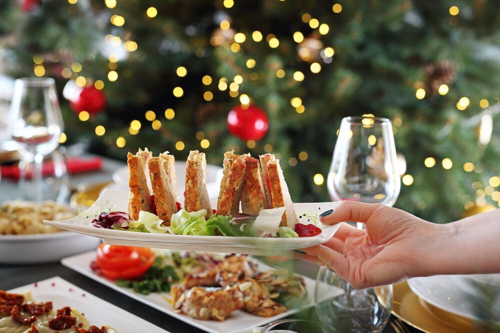 Una mano coloca un aperitivo emplatado en una mesa festiva, con luces navideñas desenfocadas al fondo