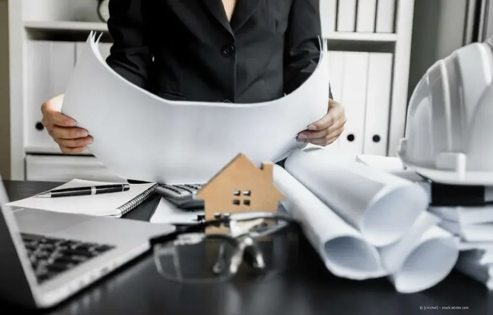 Eine Person prüft Architekturpläne an einem Schreibtisch mit Laptop, Modellhaus und zusammengerollten Plänen.
