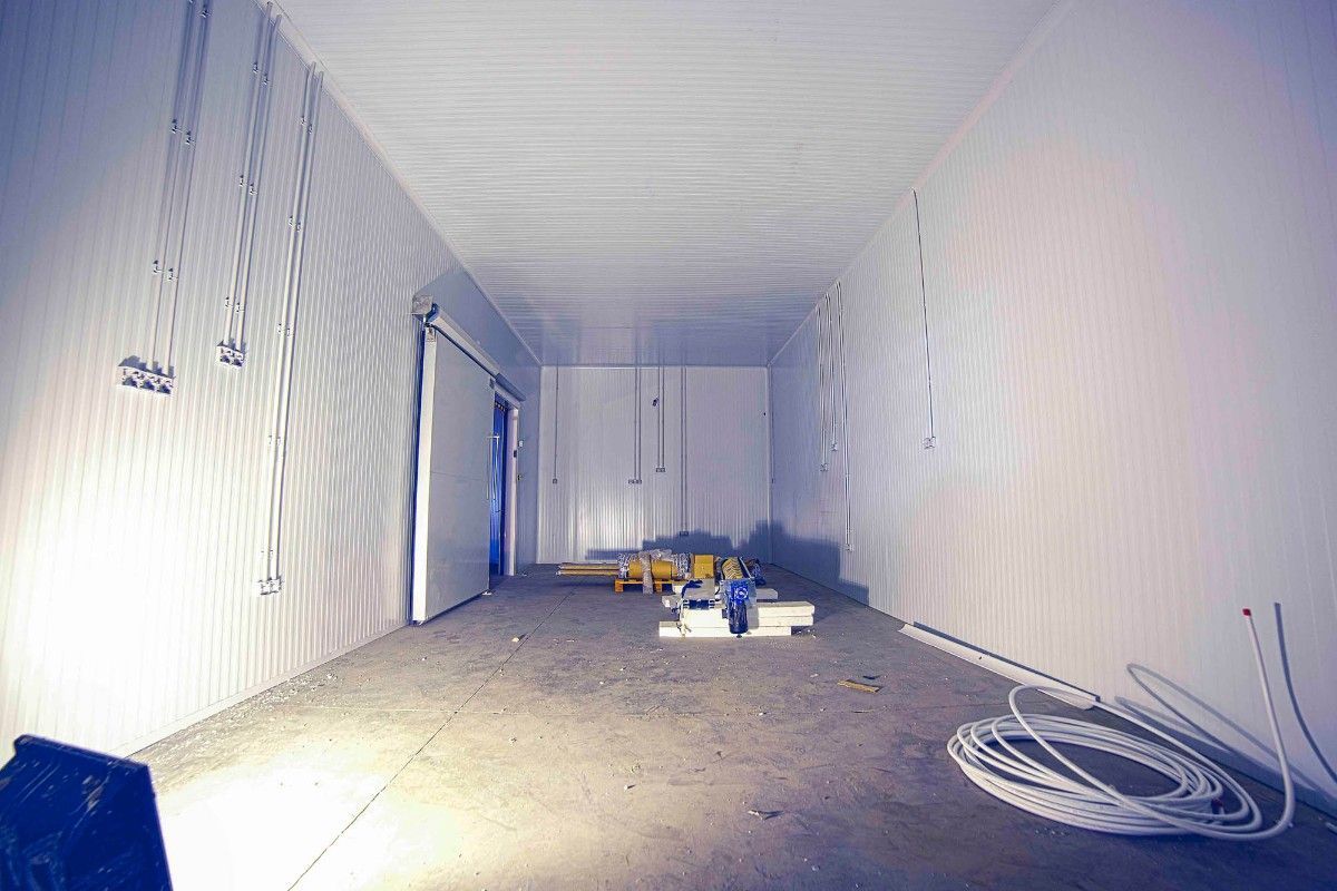 Una habitación larga y estrecha con paredes revestidas de paneles blancos, una puerta corredera a la izquierda y un cable blanco enrollado en el suelo de hormigón.