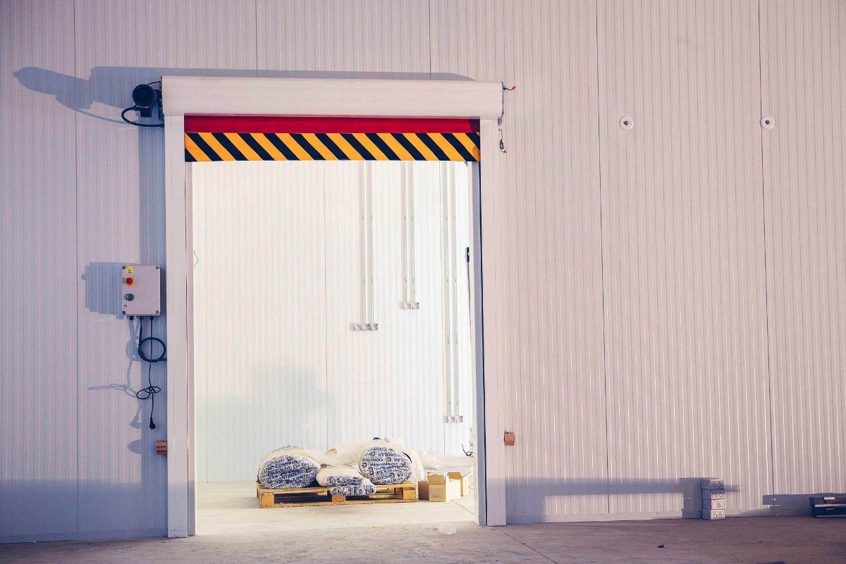 Una puerta abierta de un almacén industrial con franjas de advertencia amarillas y negras deja ver palés de mercancías en su interior.