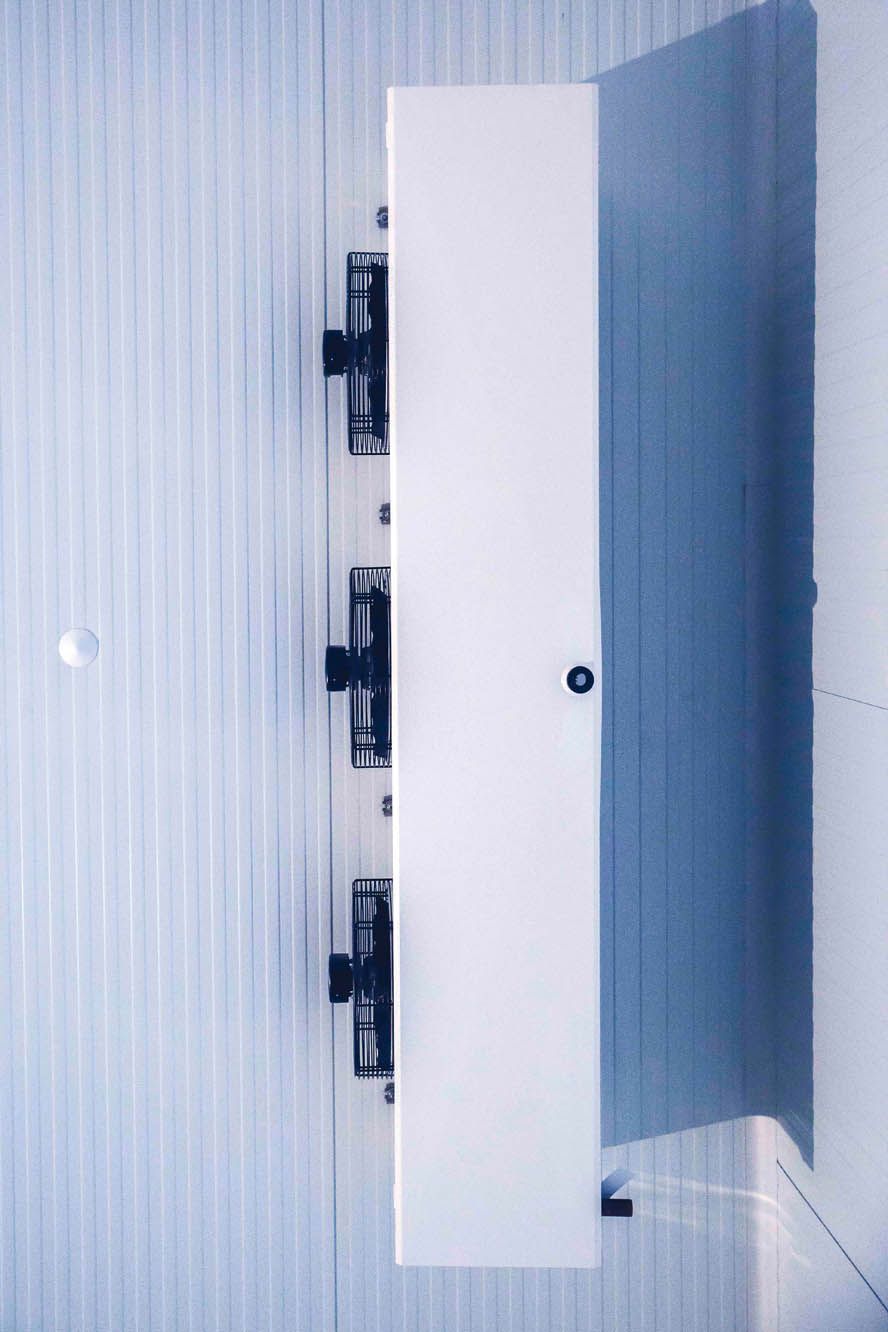 Una unidad de refrigeración comercial blanca con tres ventiladores montada sobre una pared blanca revestida verticalmente con paneles.