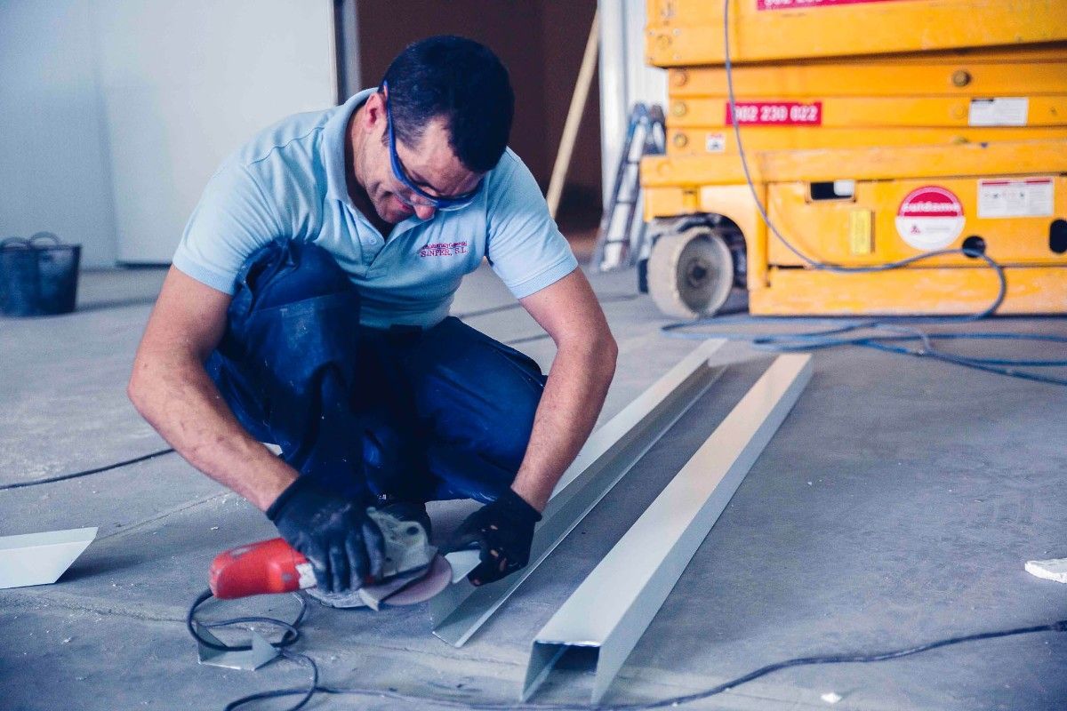 Una persona, con guantes y gafas de seguridad, está arrodillada en el suelo de un taller, utilizando una amoladora eléctrica en una pieza de canal metálico.