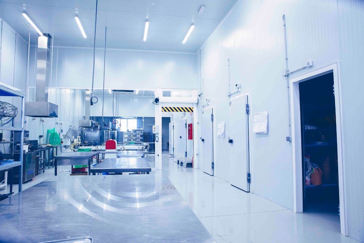 Una cocina industrial o planta de procesamiento de alimentos, bien iluminada y aséptica, con mesas de trabajo metálicas y múltiples puertas de acceso.