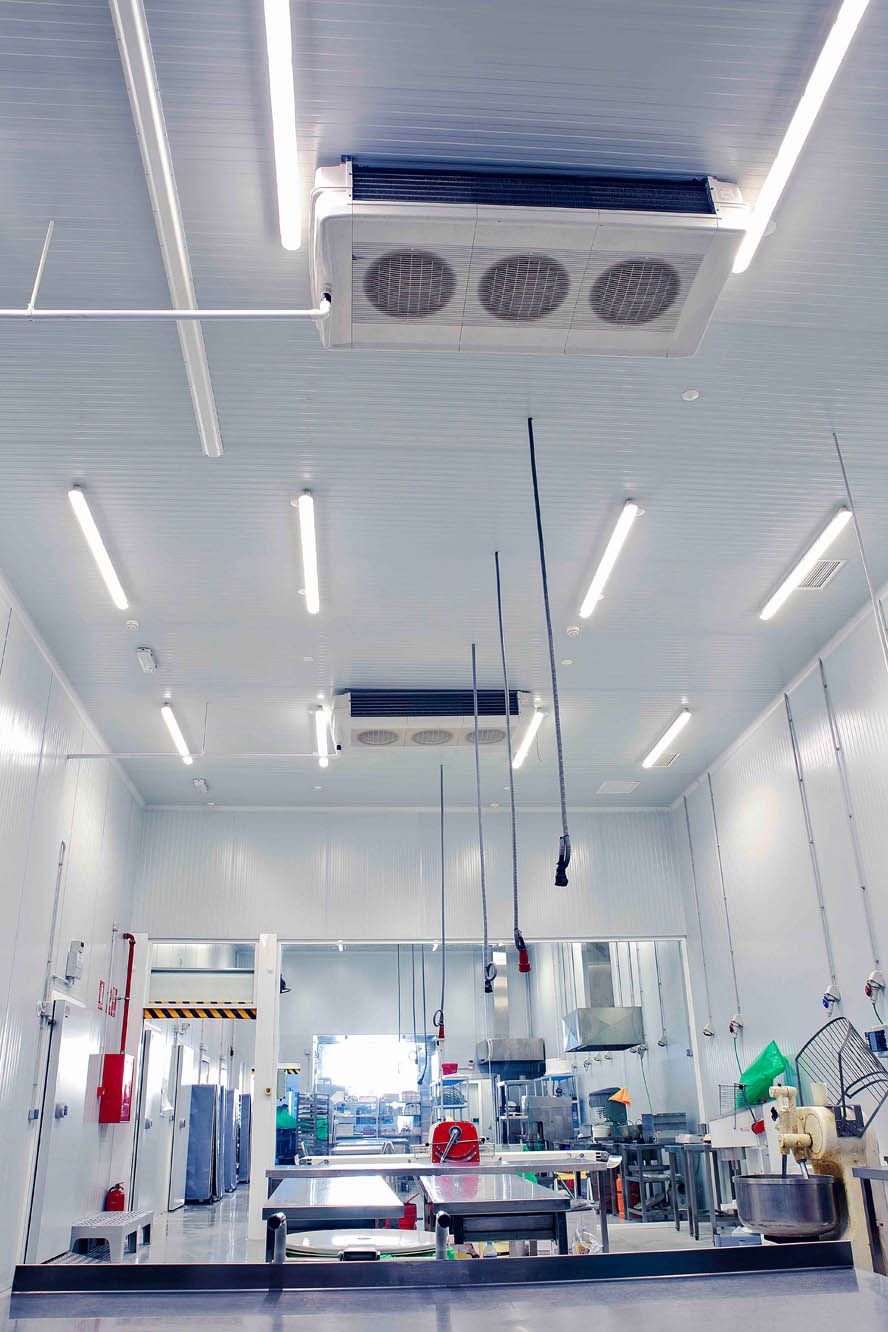 Una cocina industrial estéril o una planta de procesamiento de alimentos con paredes blancas, mesas de acero inoxidable y rejillas de ventilación en el techo.