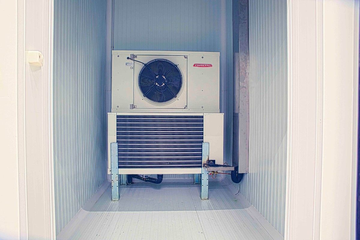 Una gran unidad de refrigeración industrial blanca, con ventilador y rejilla, se encuentra dentro de una habitación con paredes de metal corrugado de color azul claro.
