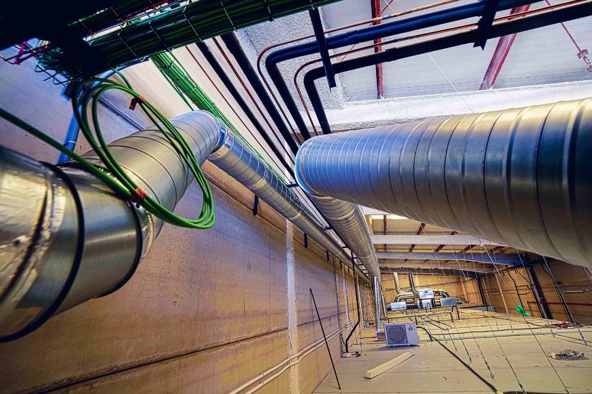 Conductos de ventilación metálicos y cables verdes colgando del techo en un gran edificio industrial sin terminar.