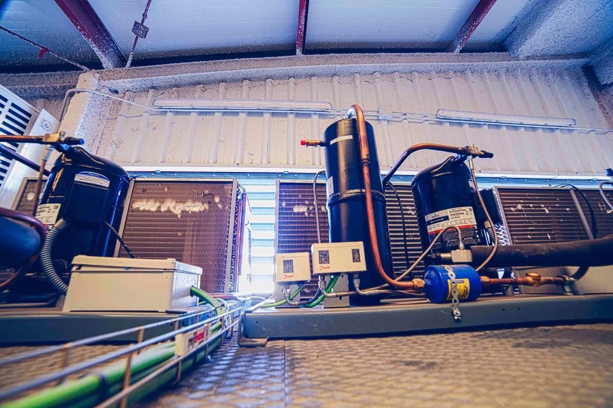 Dos compresores industriales negros y sus unidades de refrigeración asociadas se encuentran sobre un suelo metálico dentro de una instalación con techo metálico.