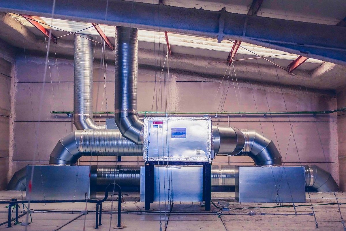Unidad de climatización industrial con conductos de ventilación metálicos instalada en una sala amplia con techos altos.