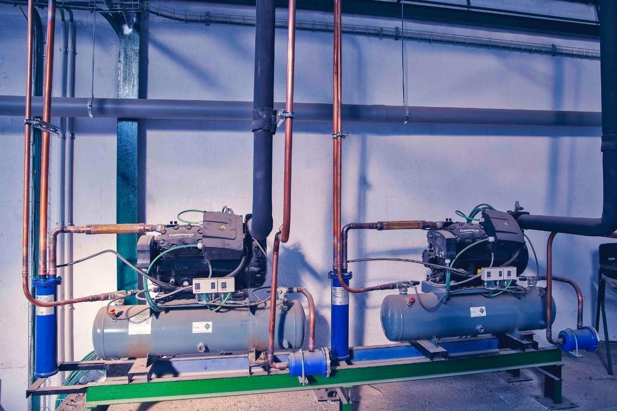Dos unidades compresoras de refrigeración industrial con tuberías de cobre y carcasas de filtro azules instaladas contra una pared blanca.