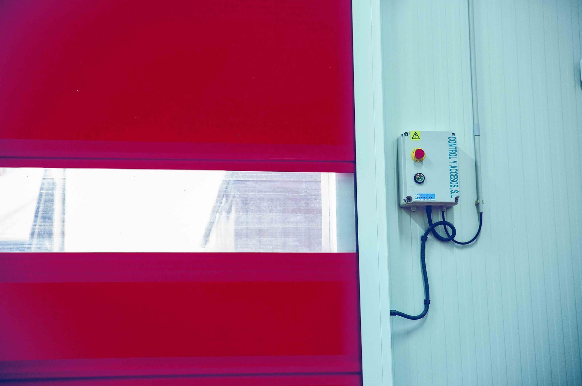 Una puerta enrollable industrial roja con una ventana transparente, situada junto a una caja de control montada en la pared con un botón de emergencia rojo.