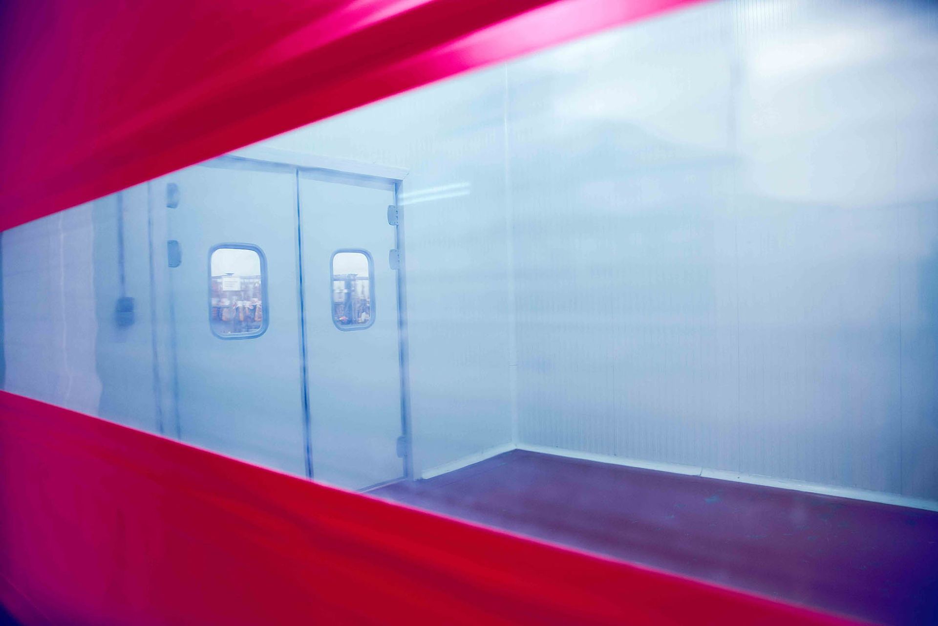 Vista a través de una ventana con borde rojo hacia una habitación limpia, luminosa y de paredes blancas con puertas dobles.