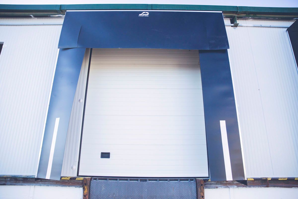Un muelle de carga con una puerta basculante blanca enmarcada por sellos de muelle negros en una pared de almacén de color claro.