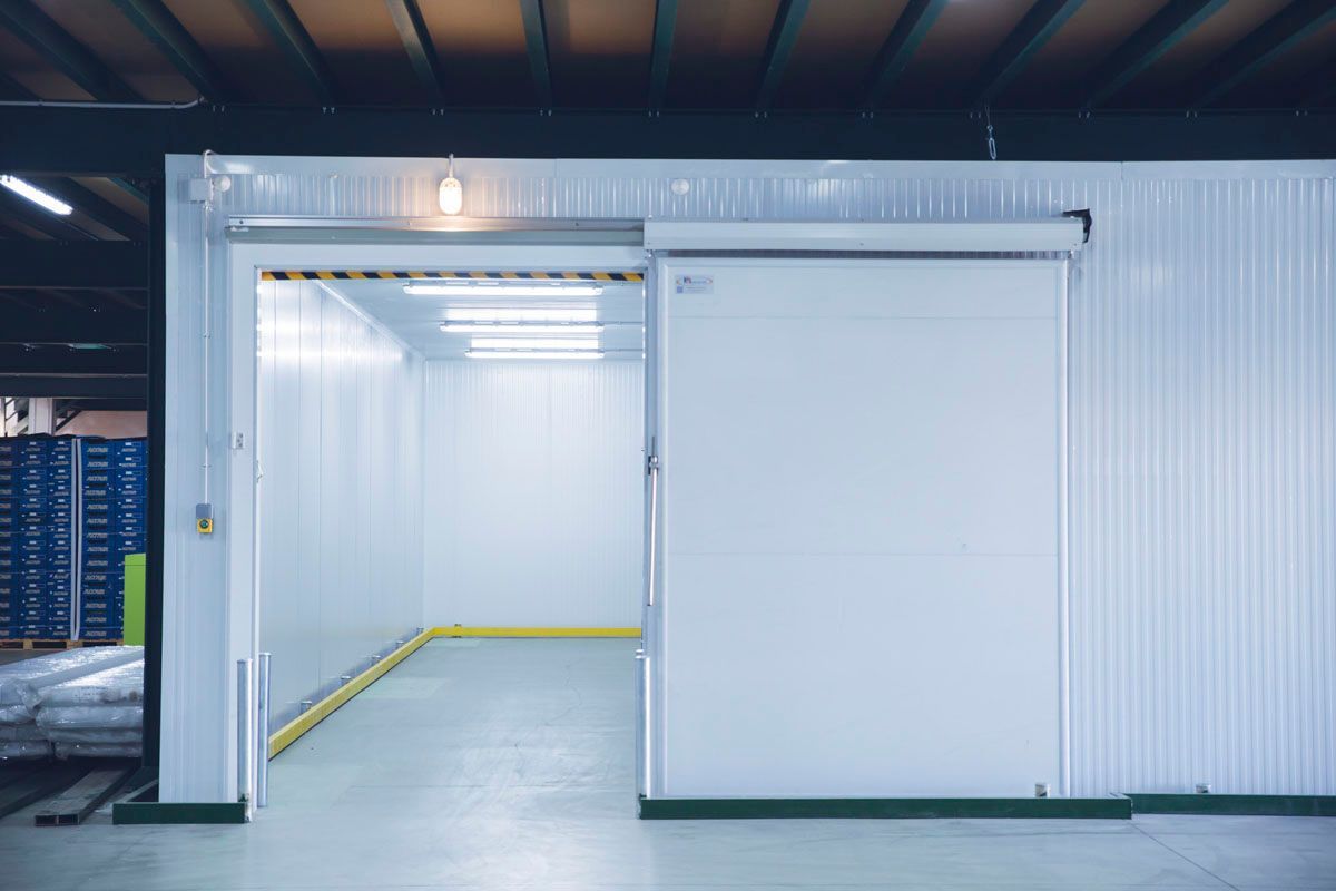 Una puerta corredera blanca abierta da acceso a un almacén industrial o cámara frigorífica vacía y bien iluminada.