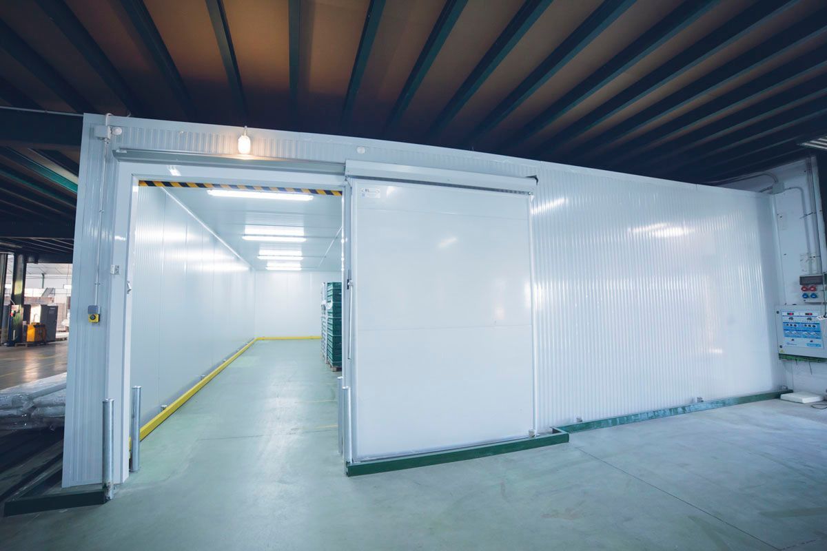 Una amplia cámara frigorífica industrial de color blanco, con puerta corredera e iluminación cenital brillante, ubicada en un entorno de almacén.