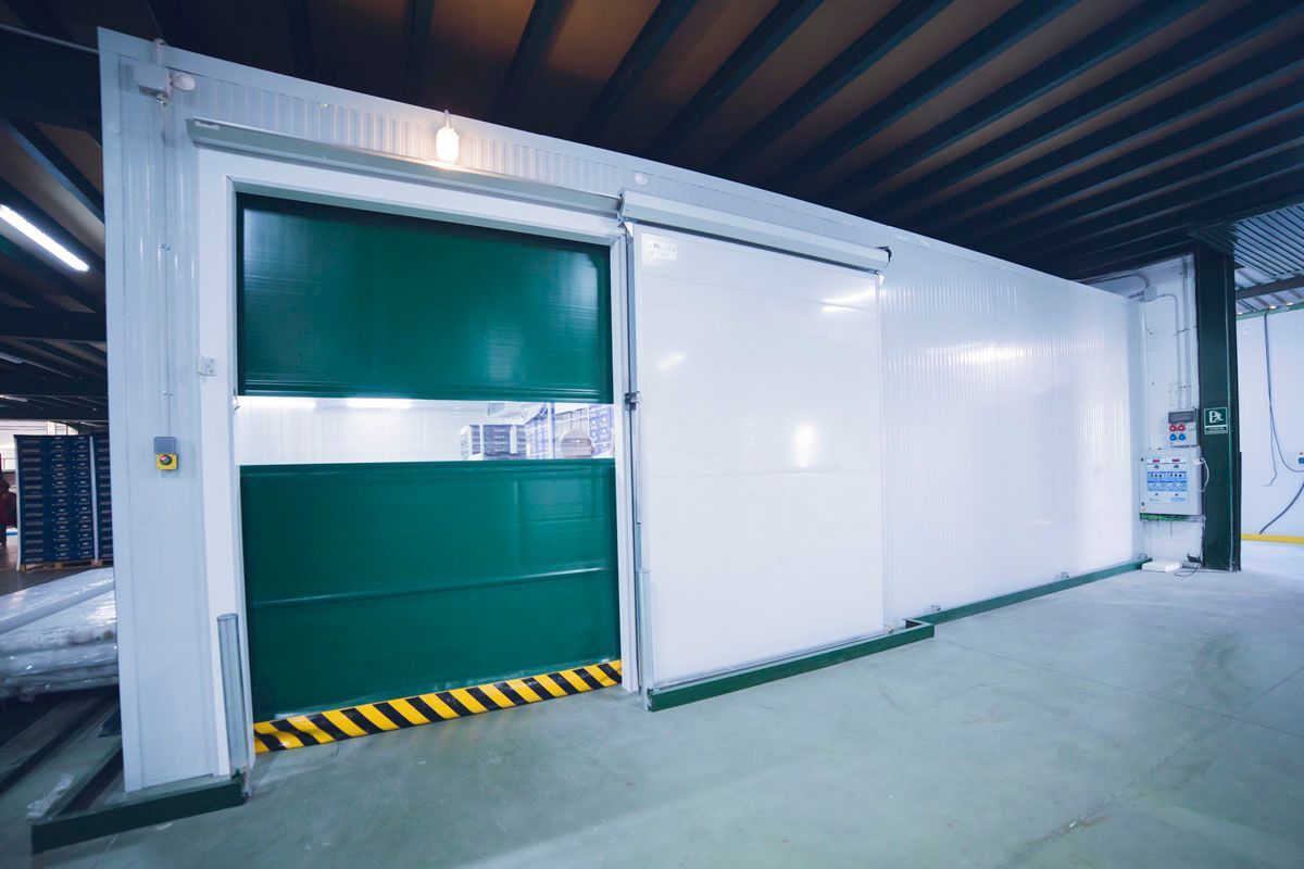 Una gran puerta corredera industrial de congelador, de color verde y blanco, con cinta de advertencia amarilla y negra en el borde inferior.