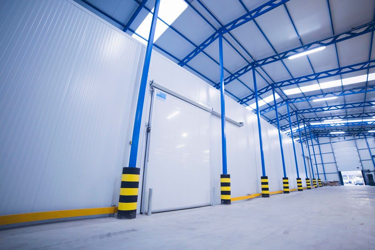 Un almacén amplio y moderno con paredes blancas, estructura metálica azul y una gran puerta corredera con marcas de seguridad amarillas.