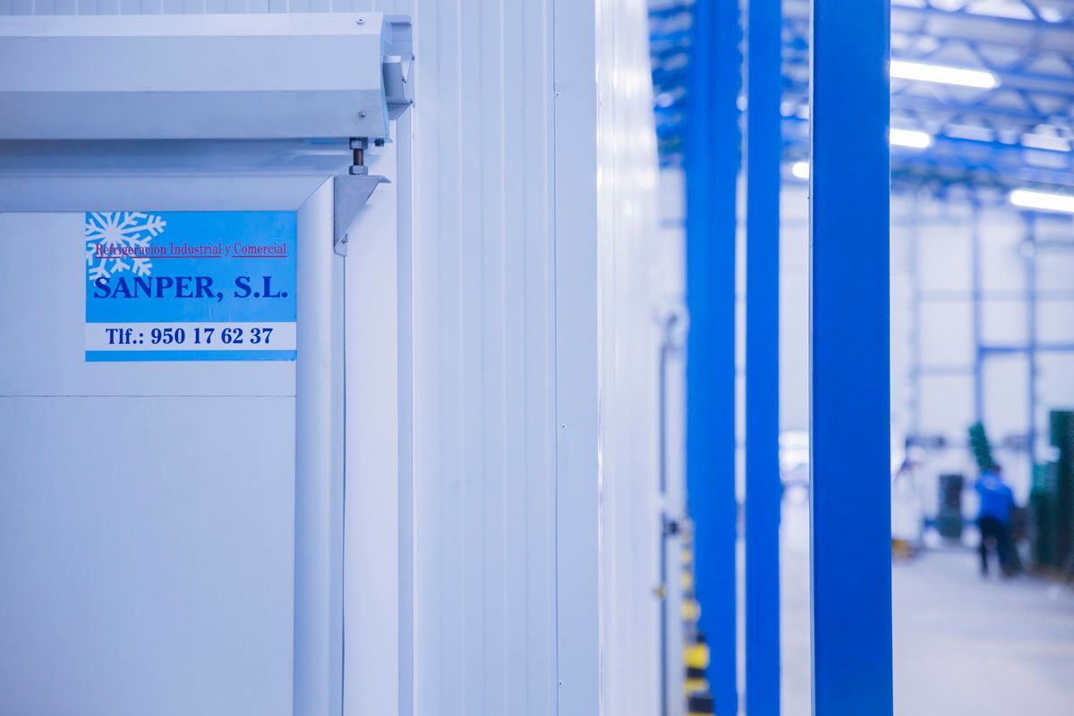 Primer plano de la puerta de un frigorífico industrial blanco con una pegatina de SANPER, SL en un almacén bien iluminado.