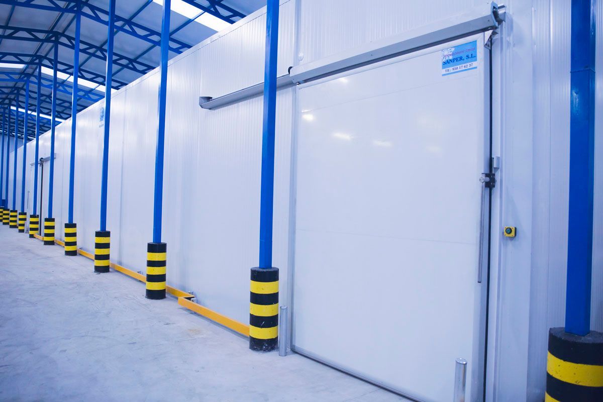 Una puerta corredera industrial blanca en una gran cámara frigorífica, ubicada en un almacén con columnas azules y barandillas de seguridad amarillas.