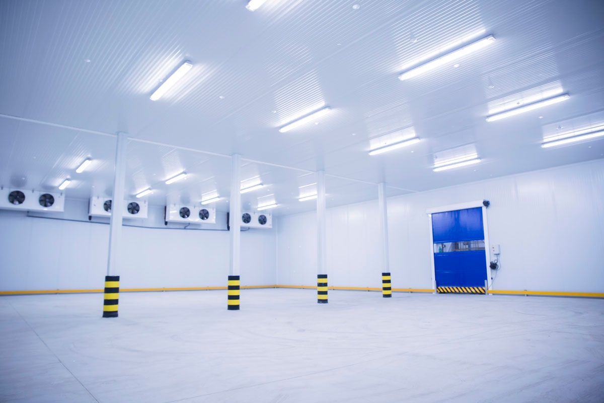 Un almacén frigorífico industrial vacío y muy iluminado, con unidades de refrigeración, pilares de soporte y una puerta azul.