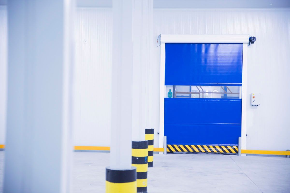 Una puerta enrollable industrial azul de alta velocidad instalada en una pared blanca con marcas de seguridad en el suelo de color amarillo y negro.