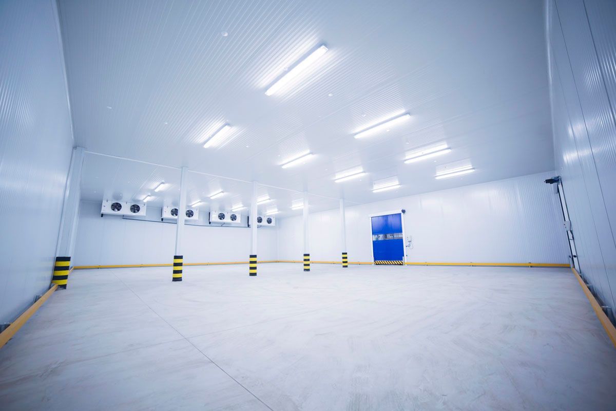 Una cámara frigorífica vacía y aséptica, con paredes blancas, luces montadas en el techo y grandes unidades de refrigeración a lo largo de una de las paredes.