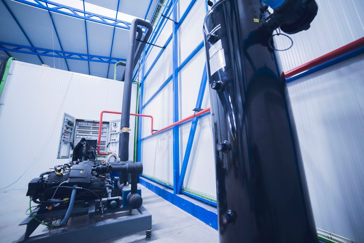 Un gran tanque vertical de color negro se alza en primer plano de una habitación que contiene maquinaria industrial y vigas de soporte azules.