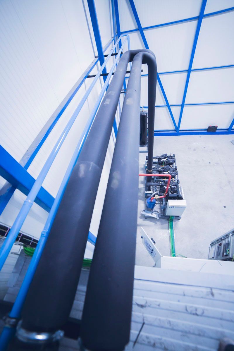 Vista desde arriba de dos grandes tuberías aisladas que discurren verticalmente junto a una estructura azul situada sobre bombas industriales.