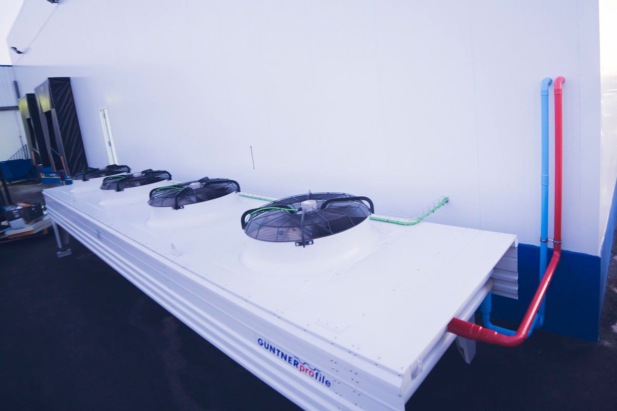 Una unidad enfriadora industrial blanca para exteriores, con cuatro ventiladores grandes, está colocada contra una pared y conectada por tuberías rojas y azules.