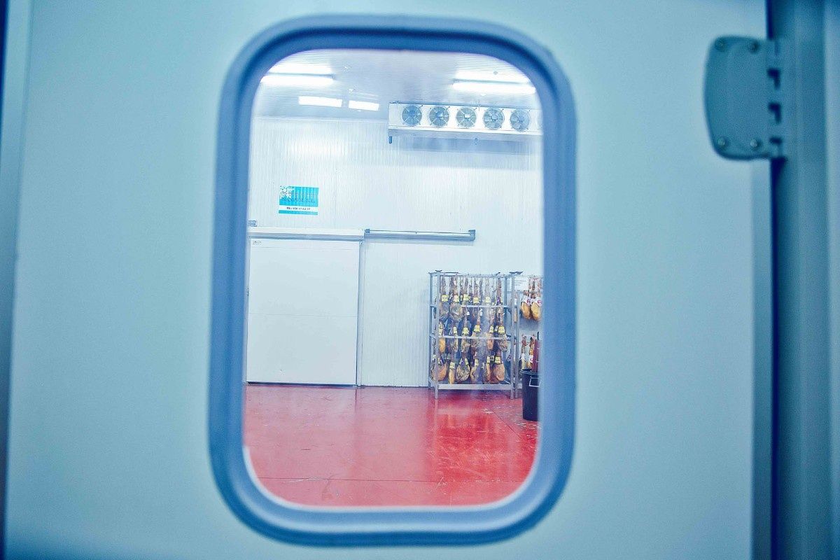 Vista a través de la ventana de una puerta hacia una cámara frigorífica para alimentos con suelos rojos, paredes blancas y jamones curados colgando.