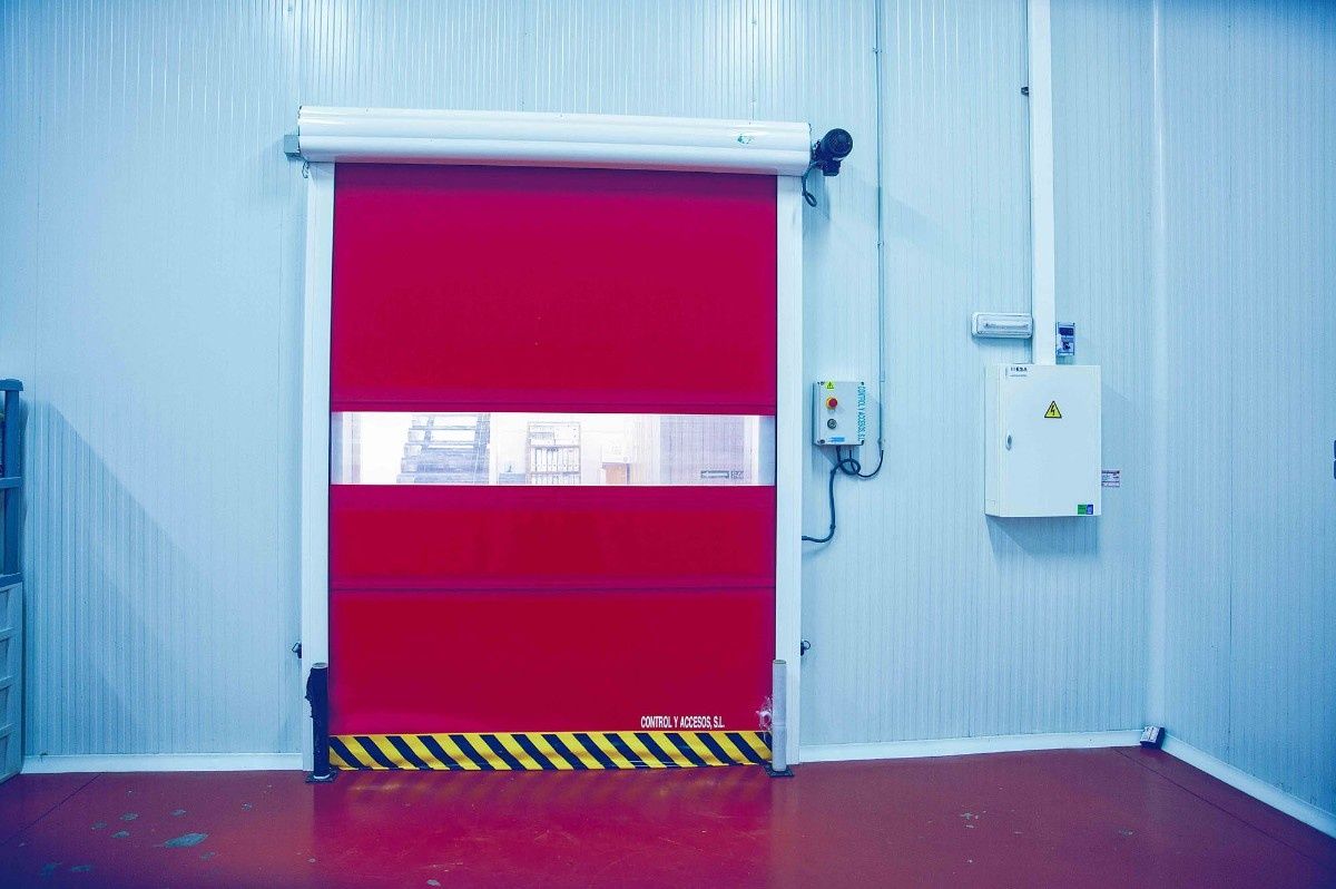 Una puerta enrollable roja de alta velocidad con un panel de visión transparente, montada sobre una pared industrial blanca encima de un suelo rojo.