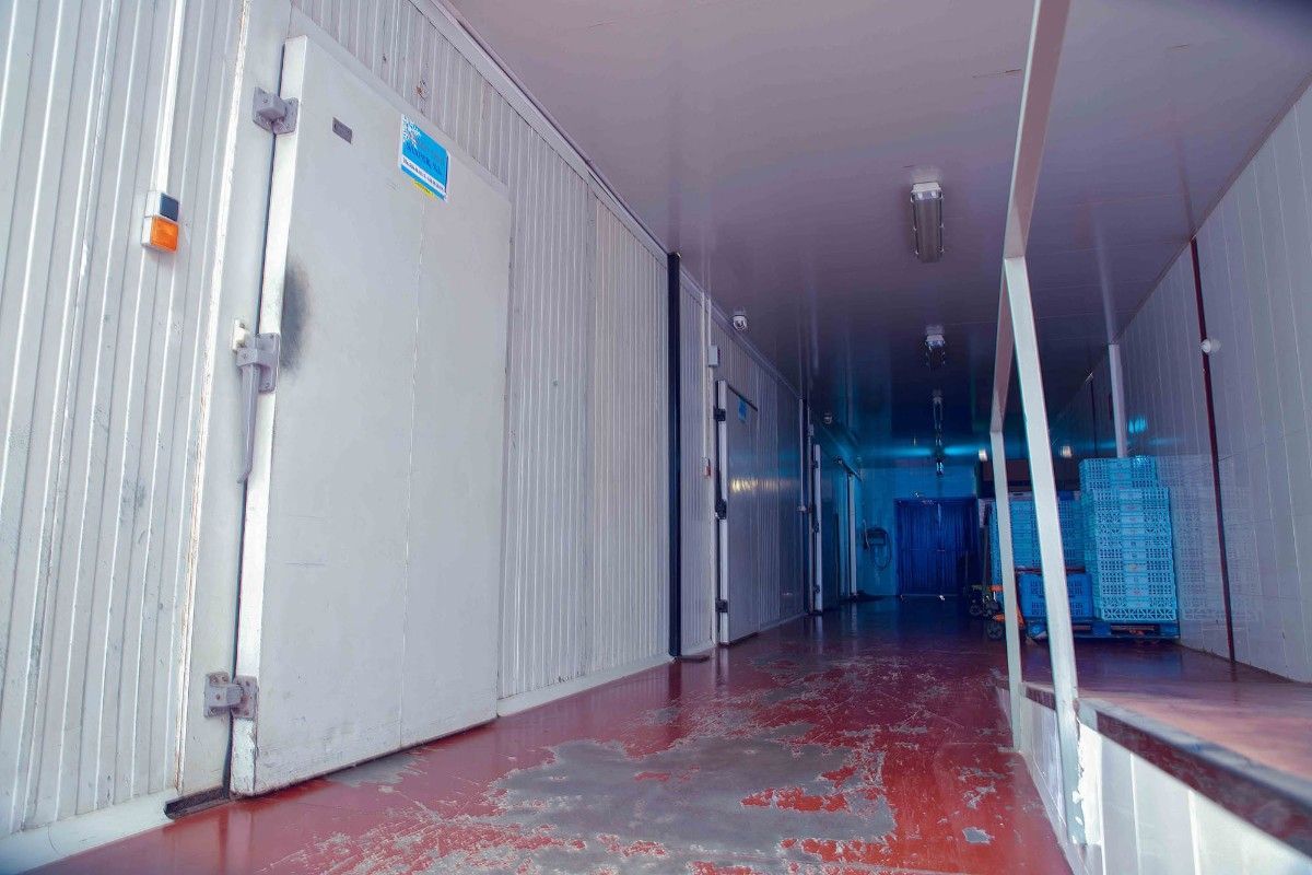 Un pasillo industrial con poca luz, con una gran puerta de frigorífico blanca a la izquierda y el suelo pintado de rojo.